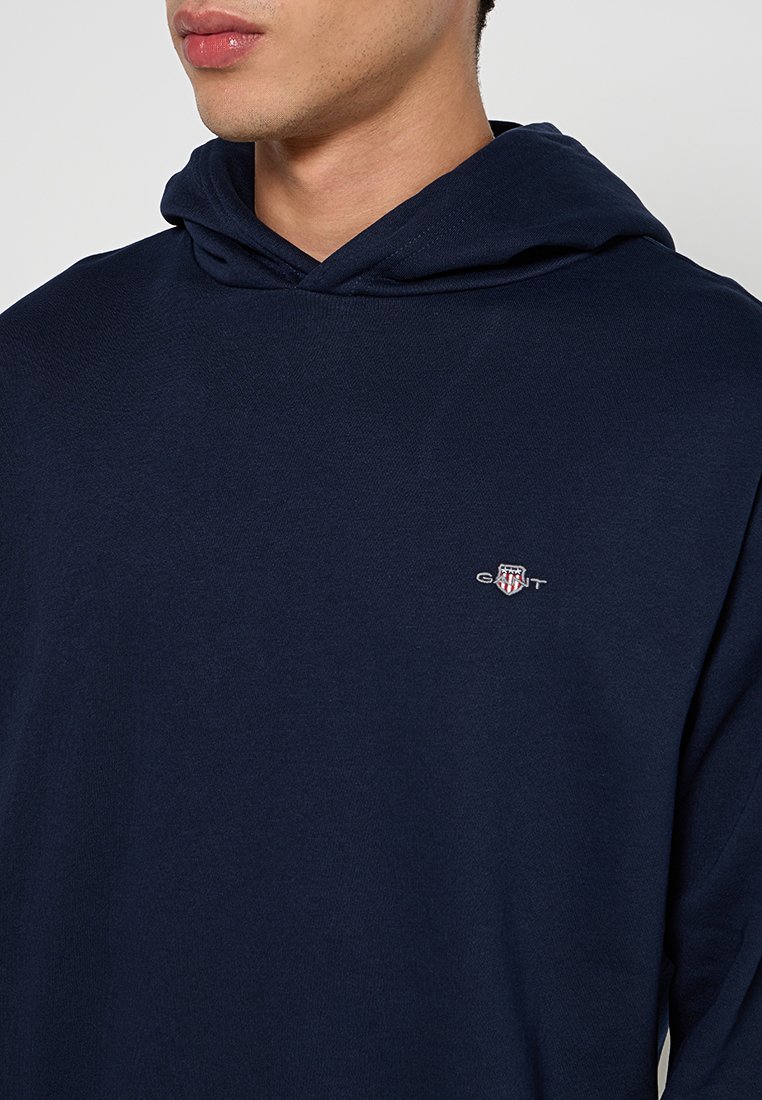 Dunkelblaues Kapuzenpullover aus einem Baumwollmischgewebe. Mit einer vorderen Kängurutasche und einem kleinen gestickten Logo auf der linken Brust.