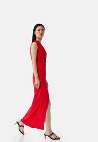 Rotes, ärmelloses Kleid mit drapiertem Ausschnitt und seitlichem Schlitz, aus glattem Stoff gefertigt. Tragen mit schwarzen Riemchenheels.