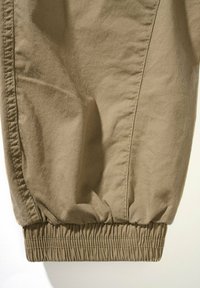 Bež hlače iz raztegljive tkanine s kavbojskimi manšetami na gležnjih, z gladko teksturo in stranskimi šivi za detajl.