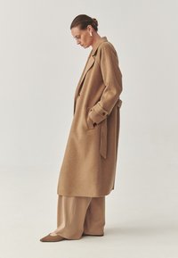 Lång tanfärgad kappa med mjuk textur, breda slag och sidofickor. Matchad med lösa beige byxor och spetsiga bruna skor.