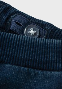 Pantaloni in denim blu scuro con una cintura a coste, dotati di un bottone in plastica trasparente e un design frontale piatto, che mostra una superficie texturizzata.