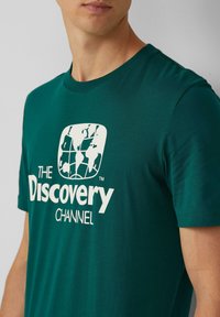 Turkusowy t-shirt bawełniany z białą grafiką Ziemi i tekstem "The Discovery Channel" napisanym pogrubioną czcionką. Krótkie rękawy, fason z okrągłym dekoltem.