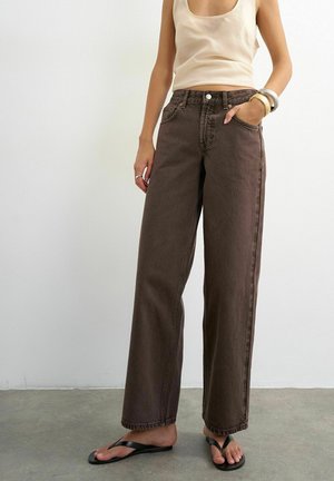 Kvinde iført beige ærmeløs top, brune højttaljede viddebukser, sorte flip-flops og armbånd står på gråt gulv.