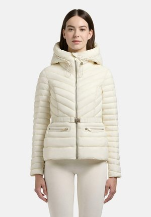 Kvinde iført en cremefarvet quiltet jakke med hætte, lynlås foran, bælte og lynlåslommer, stående mod en ensartet baggrund.