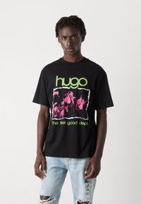Svart bomull T-skjorte med grafisk trykk i rosa og grønt. Teksten leser "hugo" og "The feel good dept." Slitte denimjeans fullfører looken.