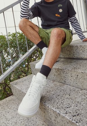 Osoba siedząca na betonowych schodach na zewnątrz, ubrana w białe sneakersy Nike, czarno-białą bluzę z długimi rękawami w paski pod czarnym t-shirtem oraz zielone krótkie spodenki.