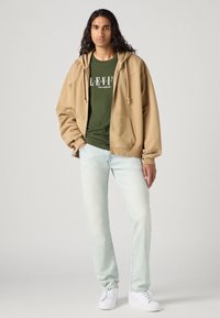 Beige hettegenser med snøring, grønn trykket t-skjorte, lyseblå jeans, og hvite joggesko. Modellen står mot en nøytral bakgrunn.