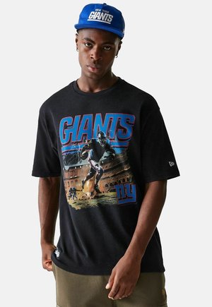 T-shirt oversize nera con una grafica colorata "GIANTS" e un design di un giocatore di football. Abbinato a un cappellino blu con la scritta "NEW YORK GIANTS."