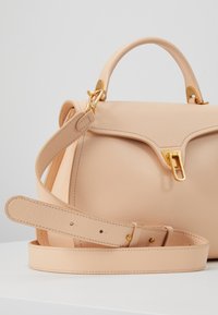 Beige läderhandväska med ett handtag upptill, justerbar axelrem och klämlås i guldton på en vit yta.