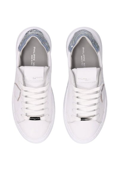Zapatillas de cuero blanco con textura suave, que presentan un acento metálico, diseño con cordones y un ribete de purpurina azul alrededor de la abertura.