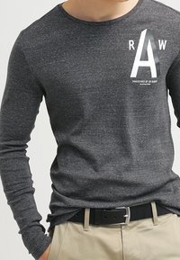 Mörkt grå långärmad T-shirt i mjukt tyg, med en vit grafisk bokstav "A" och text på vänster bröst.