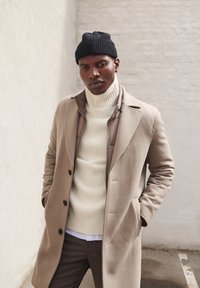Beige wollen jas met een getailleerd ontwerp, gedragen over een crèmekleurige geribde coltrui. Zwarte gebreide beanie voegt textuur toe; grijze achtergrond.