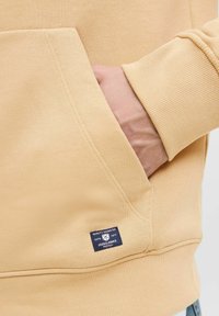 Sudadera de color amarillo claro con un bolsillo tipo canguro y puños acanalados, con una pequeña etiqueta azul en el bolsillo que muestra los detalles de la marca.