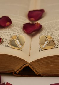 Guldörhängen i hjärtform med en central diamantskärva, placerade på en öppen bok omgiven av rosenblad. Slät yta, polerad finish.