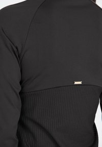 Schwarzes Sporttop mit strukturierter Rippoptik im unteren Bereich, glattem Stoff im oberen Bereich und einem kleinen metallischen Logoakzent.