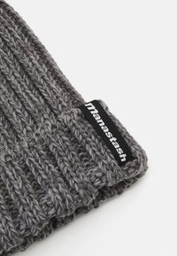 Grå strikket lue med ribbestruktur og svart merkelapp med merkevarenavnet "manastash." Stoffet ser tykt og varmt ut.