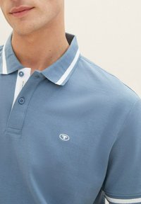 Ljusblå polo skjorta gjord av texturerad tyg, med en vit krage med randiga detaljer och en logotyp på vänster bröst. Två-knapprad.