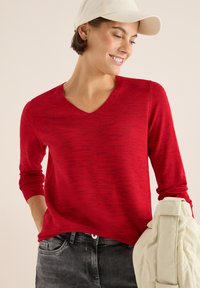 Maglione rosso a maniche lunghe con scollo a V e un motivo testurizzato, abbinato a jeans grigio scuro e una giacca color crema.