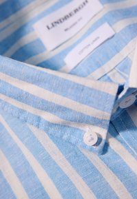Camicia in lino a righe blu chiaro e bianche con colletto button-down e etichette in stoffa che indicano materiale in cotone al 100%.