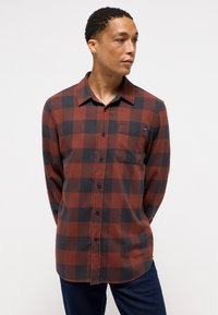 Rød og sort ternet flannel skjorte med lange ærmer, knaplukning foran, enkelt brystlomme og et marineblåt mærke. Båret med mørke jeans.