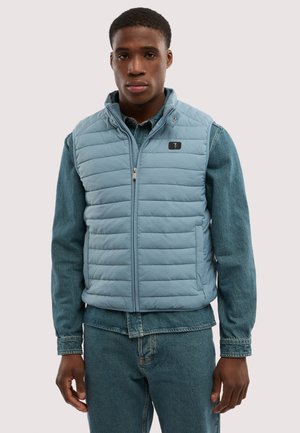 Giovane uomo che indossa un gilet trapuntato azzurro chiaro sopra una camicia di jeans e jeans, in piedi con un'espressione neutra contro uno sfondo semplice.