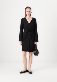 Must musträtik, millel on pikad varrukad, V-kaelus ja esine sõlmikas. Sile kangas, põlvikukõrgus, koos musta käekotiga ja lapikute kingadega.