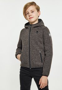 Grå zip-up huvtröja med en strukturväv, utrustad med huva, två dragkedjor och en logotyp på vänster ärm. Bärs över svarta byxor.