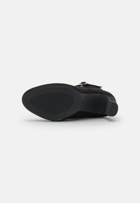 Zapato de ante negro con suela de goma texturizada, que presenta una puntera redondeada y una correa con herrajes en oro rosa. La suela tiene un patrón acanalado.