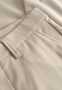 Kohevpildis beežist tekstuuriga kangast püksid, keskendudes vöö ja vööaasade detailidele.