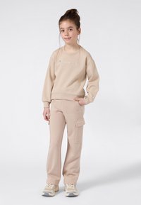Fille jeune se tenant avec une main dans la poche, portant un sweat à capuche beige de Champion, un pantalon cargo assorti et des baskets beiges sur fond uni.