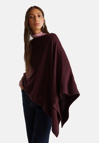 Donna che indossa un poncho in maglia asimmetrico di colore viola scuro sopra un dolcevita viola chiaro e jeans scuri, in piedi con una mano in tasca.