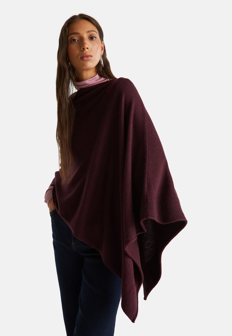 Donna che indossa un poncho in maglia asimmetrico di colore viola scuro sopra un dolcevita viola chiaro e jeans scuri, in piedi con una mano in tasca.