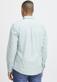 Camisa de manga larga azul claro con cuello abotonado y pliegue trasero sencillo. Hecha de una tela suave, presenta puños abotonados y aberturas laterales.