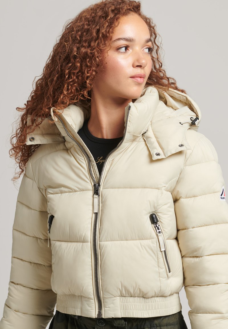 Giacca imbottita beige chiaro con colletto alto, chiusura frontale con zip e due tasche con zip. Presenta polsini e orlo elasticizzati, con un accento del logo.