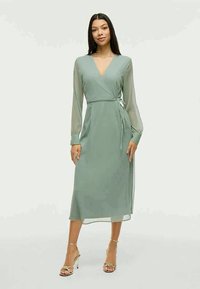 Chiffon omslagskjole i myk grønn, med lange gjennomsiktige ermer, V-hals og knyting i midjen, som når til midten av leggen.