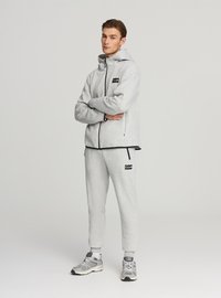 Felpa con cappuccio grigia con zip e pantaloni della tuta realizzati in tessuto morbido, con dettagli neri e logo. Modello in piedi con le braccia incrociate. Scarpe sportive.