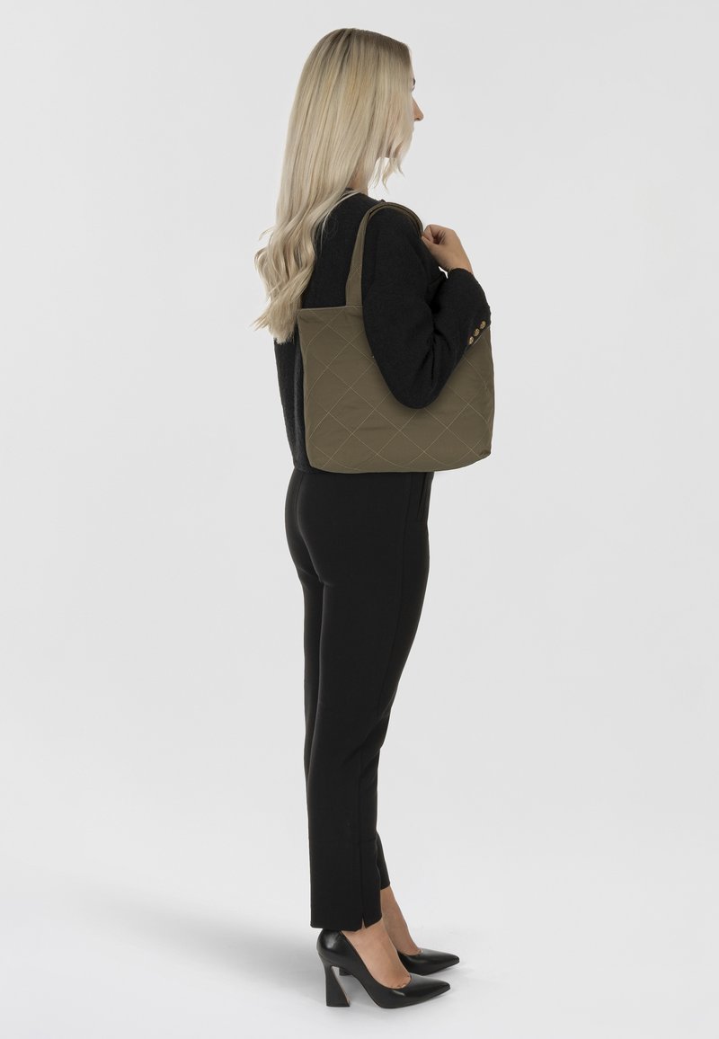 Borsa a spalla trapuntata verde oliva con forma strutturata, doppio manico e superficie testurizzata, abbinata a un outfit nero e tacchi alti.