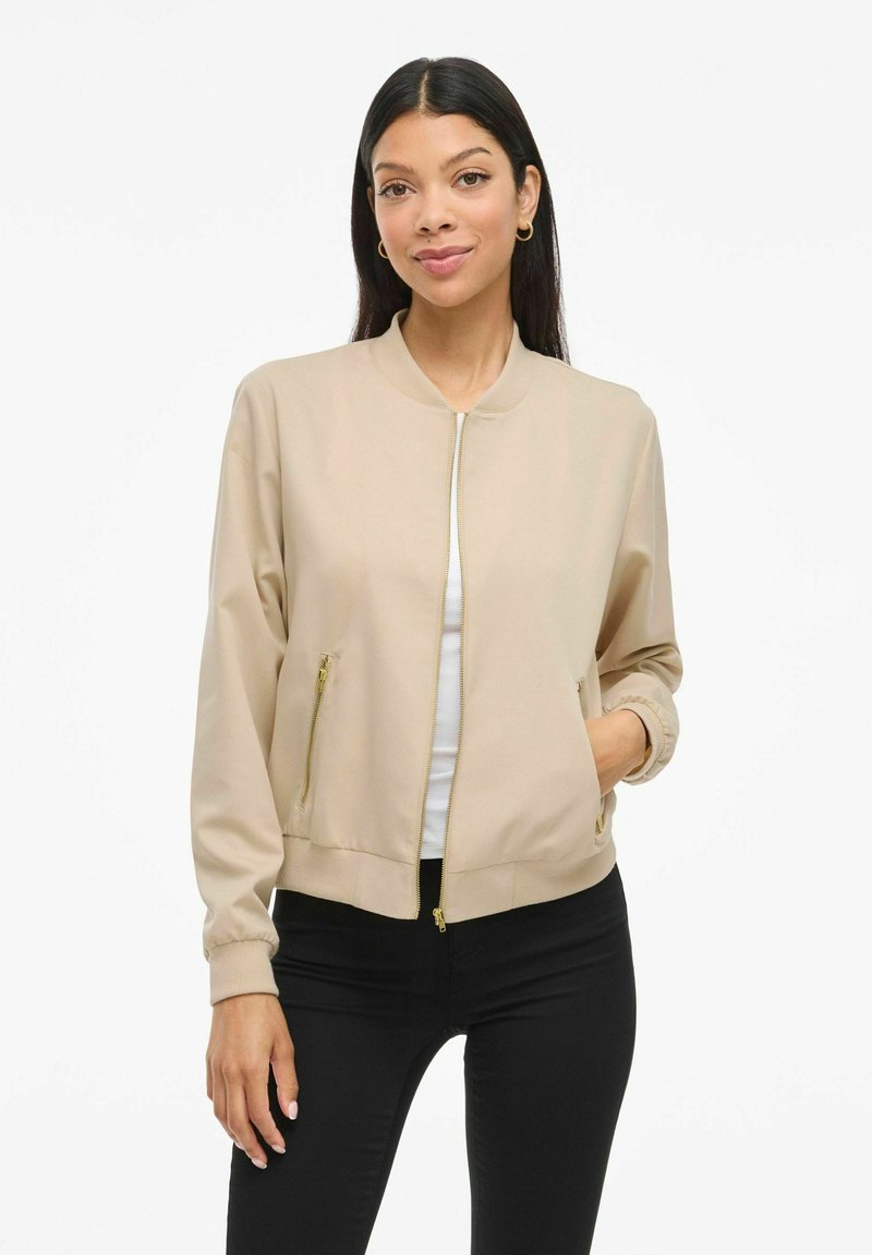 Femme aux longs cheveux noirs, portant une veste beige zippée avec poches, un haut blanc en dessous et un pantalon noir, se tenant devant un fond blanc uni.