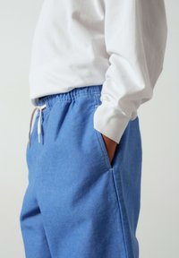 Personne portant un pantalon bleu clair à cordon et une chemise blanche à manches longues, une main dans la poche, sur un fond uni.