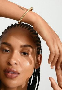 Bracciale rigido in oro indossato su un polso, con una finitura liscia e lucida. Lo sfondo è neutro, evidenziando i dettagli del bracciale.