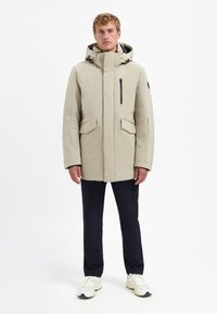 Beige geïsoleerde parka met een hoge kraag, capuchon en voorzakken met rits. De stof lijkt glad en wordt gecombineerd met donkere broek en sneakers.