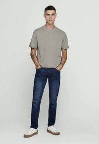 T-shirt cinza de mangas curtas emparelhada com jeans azul escuro. O modelo usa ténis brancos com solas castanhas claras. Tatuagens visíveis nos braços.