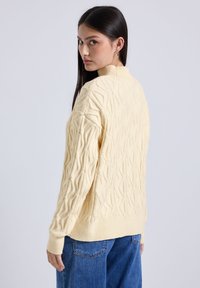 Pull en tricot crème, avec un motif de vagues texturé, col montant, coupe ample et avec des poignets et un ourlet côtelés, associé à un jean bleu.