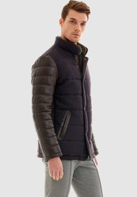 Puffjacka med en blå tweedkropp, bruna vadderade lädersleeves, hög krage och framfickor. Strukturerad tyg med släta detaljer.