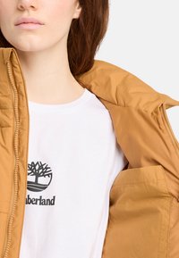 Chaqueta de plumas marrón con cremallera en la parte delantera, llevada sobre una camiseta blanca con un logo de árbol negro. Textura suave y sin patrones visibles.