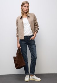 Kobieta w beżowej kurtce, białej koszuli, ciemnych dżinsach, białych trampkach, trzymająca brązową zamszową torbę na tle gładkiej jasnej ściany.