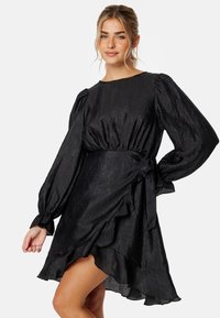 Robe noire en tissu texturé avec de longues manches bouffantes, une taille cintrée et un ourlet à volants. Détail noué sur le côté.