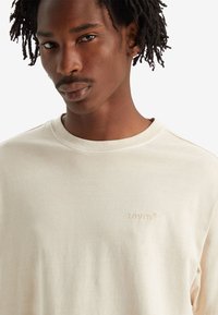 T-shirt à manches courtes de couleur beige clair en coton doux, doté d'un logo en relief discret sur la poitrine et d'un col rond classique.
