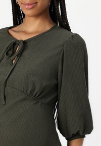 Vestido texturizado en verde oscuro con escote en forma de llave y detalle de lazo. Mangas abullonadas con puños elásticos. Tela suave con un patrón sutil.