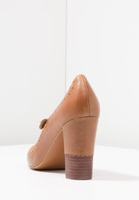 Chaussure à talon haut en cuir marron avec une pointe arrondie, un bord festonné et un talon à texture boisée, avec un détail de bride latérale.